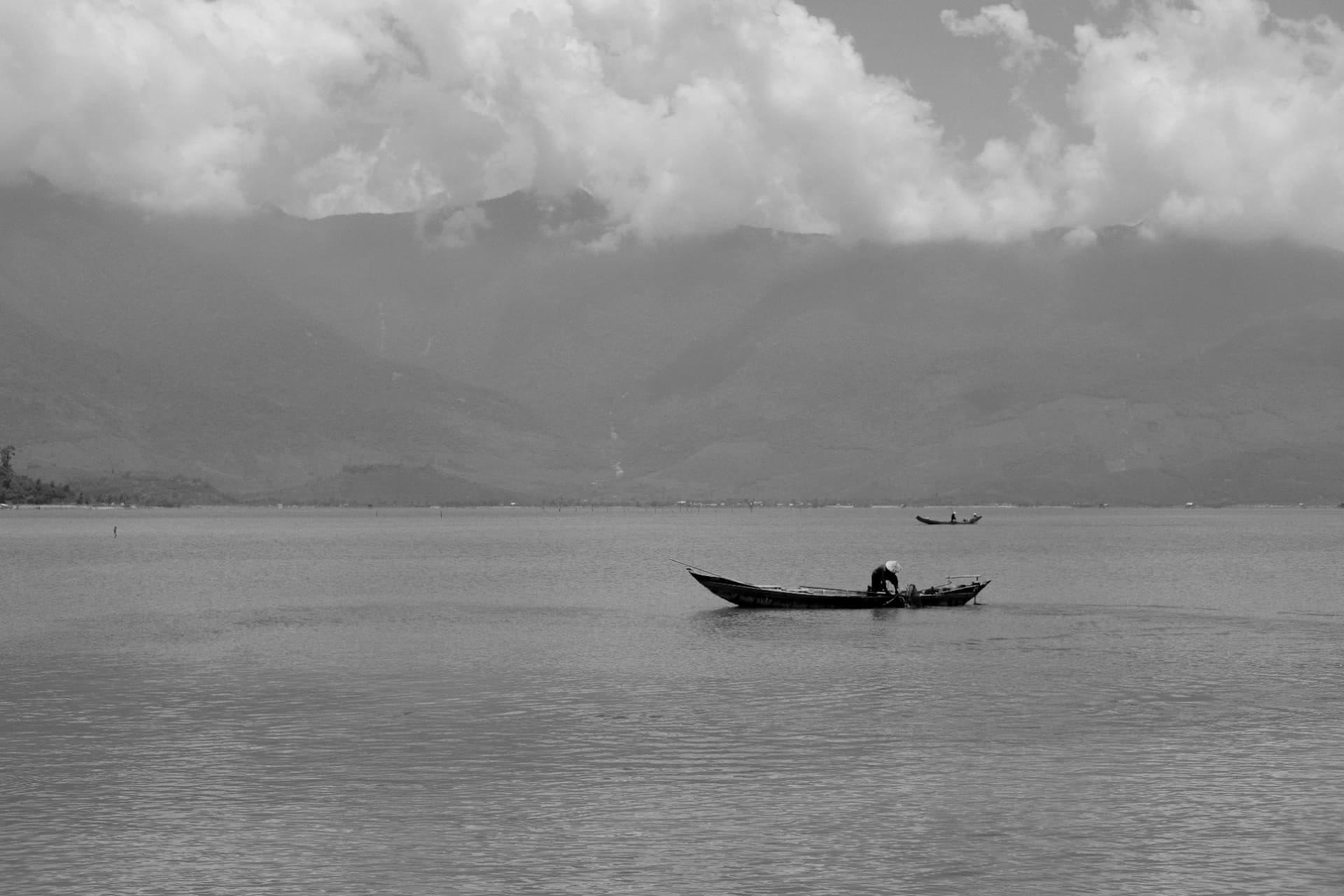 Fisherman tending nets from a wooden boat on a wide lagoon below cloud-covered mountains