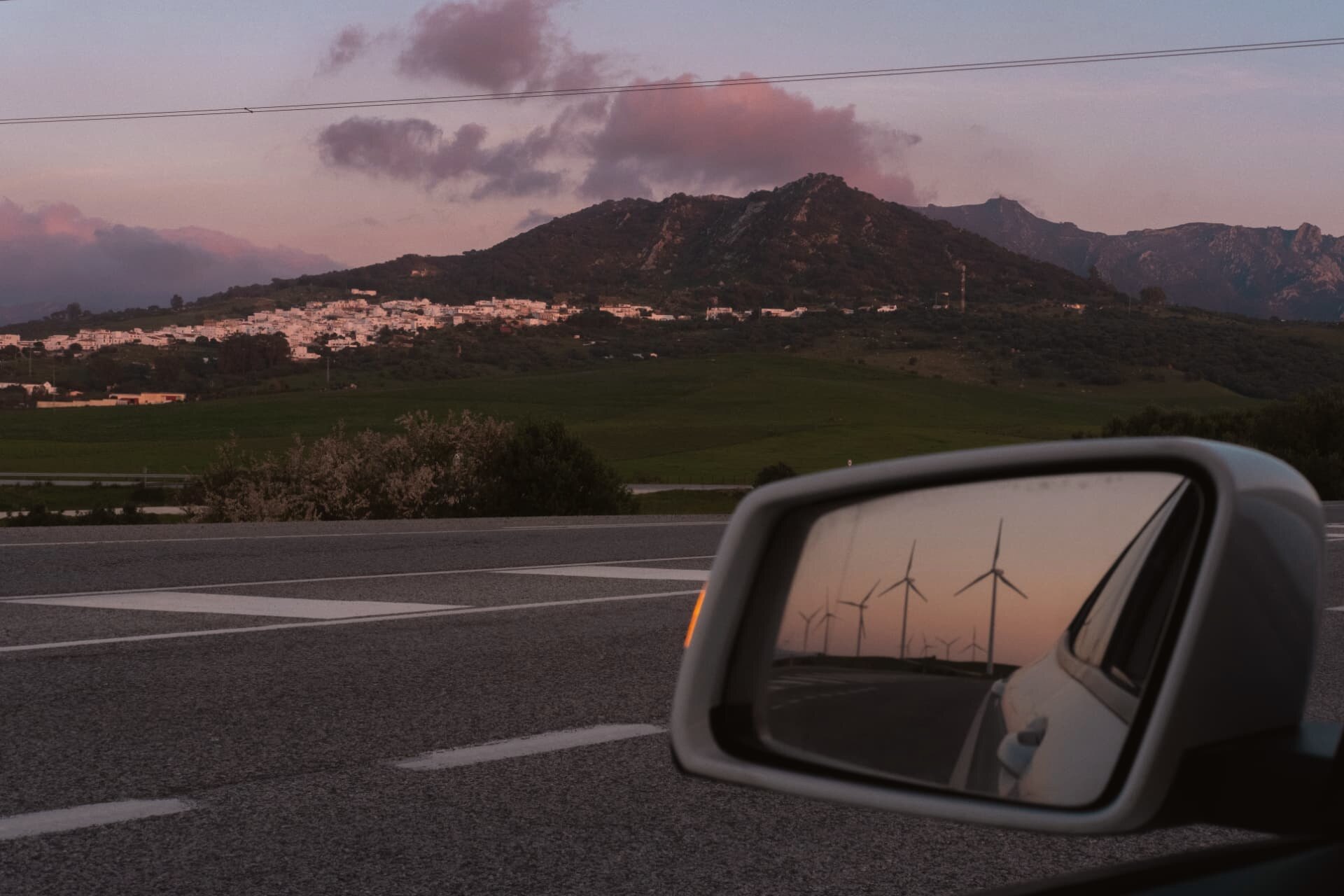 Hilltop village at dusk seen from the roadside, wind turbines reflected in the wing mirror
